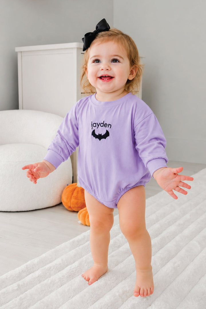 A toddler girl wearing a light purple long-sleeve romper with 'jayden' and a bat graphic printed on the chest, standing on a white textured rug in a nursery.