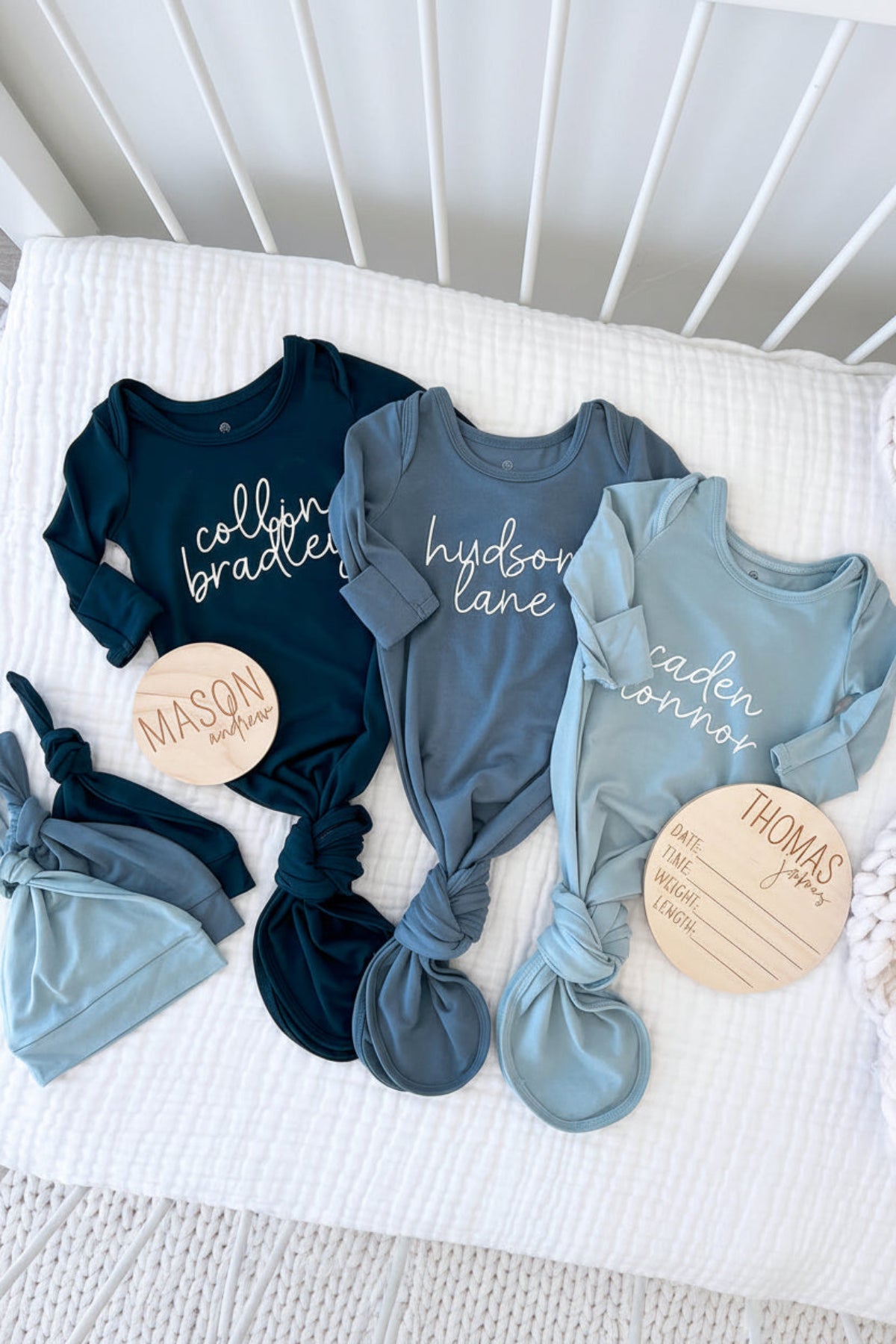 Three Caden Lane personalized knot gowns in navy, slate blue, and light blue with "Collin Bradley," "Hudson Lane," and "Caden Connor" in cursive script, paired with wooden name and birth stat signs.