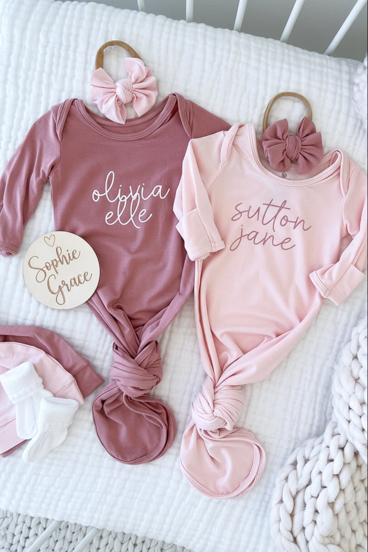 Two Caden Lane personalized baby knot gowns in mauve and blush pink with cursive names "Olivia Elle" and "Sutton Jane," styled with matching bow headbands and a wooden name sign.