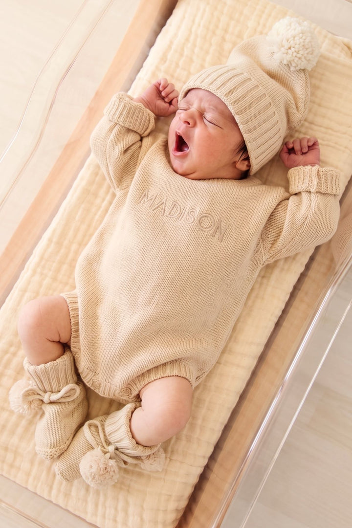 A sleeping newborn baby wearing a cream-colored knit romper, matching beanie hat with pom-pom, and booties, lying on a textured cream muslin blanket in a wooden bassinet.