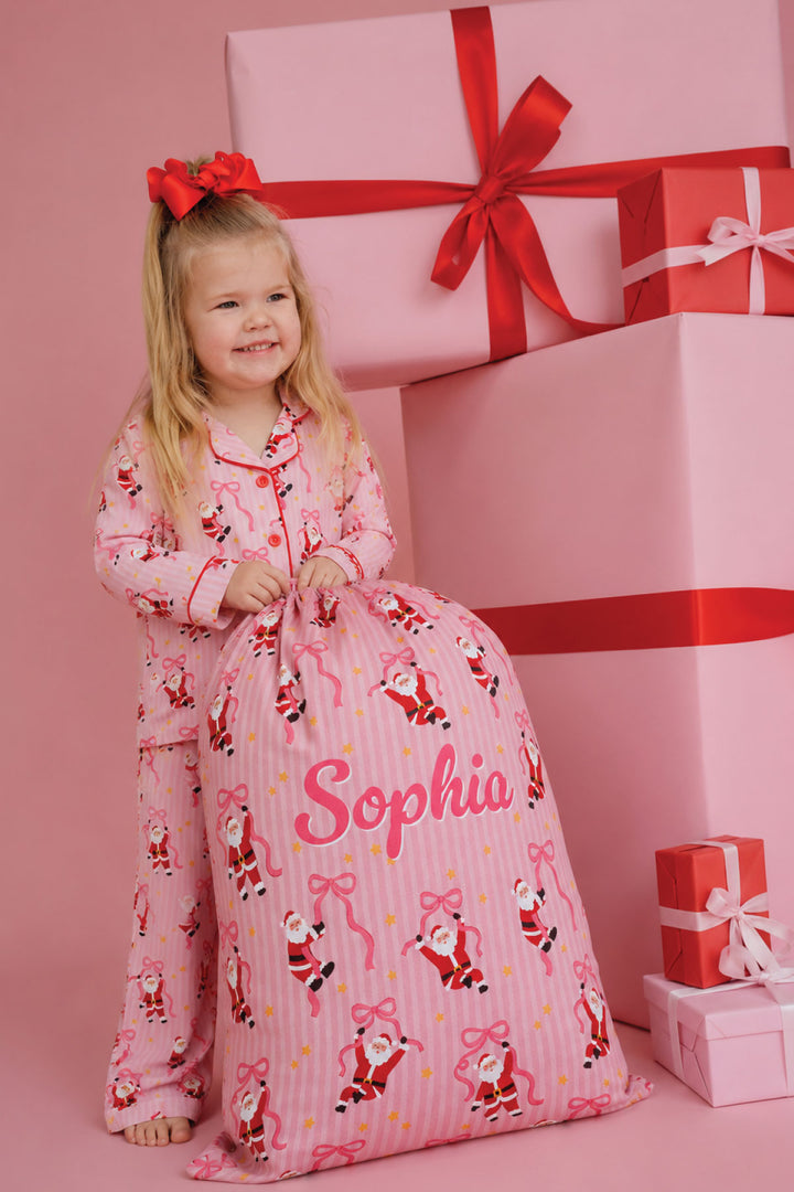 Child wearing Caden Lane Jolly Ribbons Christmas pajamas holding a personalized Christmas Santa Sack, matching festive holiday pajamas, personalized gifts, and Christmas accessories