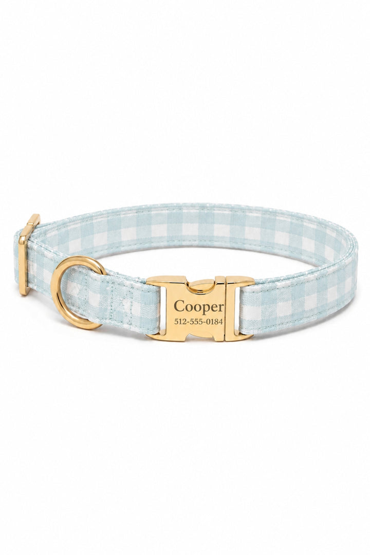 A personalized Lane & Leash dog collar featuring a light blue and white gingham check pattern, a gold-tone metal buckle engraved with the name 'Cooper' and a phone number, and a gold-tone D-ring, shown on a plain white background.