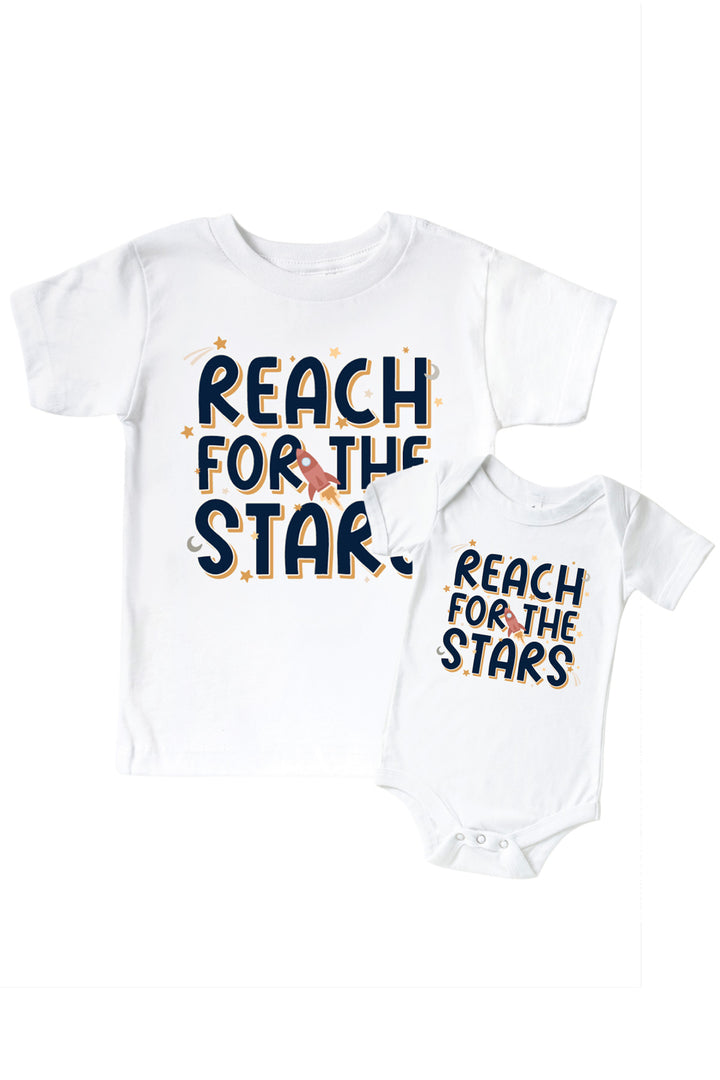 Flat lay of a matching Caden Lane white kids’ tee and bodysuit pair with a Stellar Snuggles graphic featuring a rocket, reading “Reach for the Stars.”