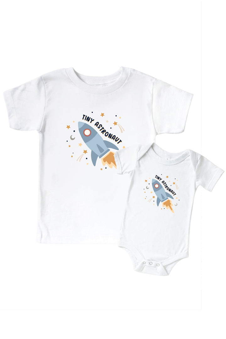 Flat lay of a matching Caden Lane white kids’ tee and bodysuit pair with a Stellar Snuggles graphic featuring a blue rocket, reading “Tiny Astronaut.”
