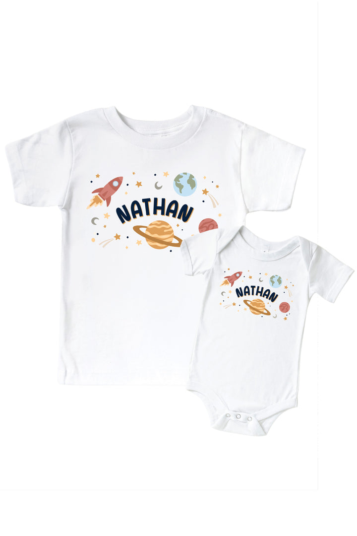 Flat lay of a matching Caden Lane white kids’ tee and bodysuit pair with a Stellar Snuggles Planets graphic featuring labeled planets and rockets.