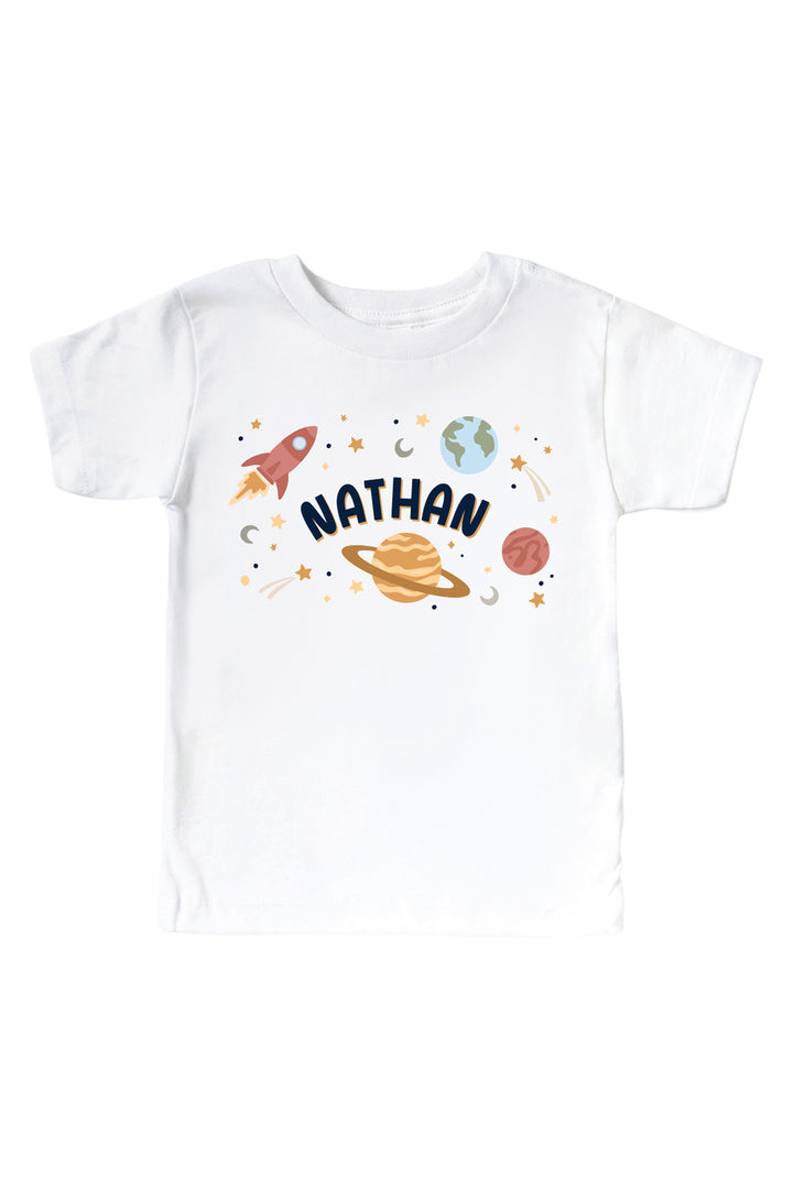 Flat lay of a Caden Lane white kids’ tee with a Stellar Snuggles Planets graphic featuring Saturn, Earth, Neptune, Mercury, Venus, and Mars with rockets.