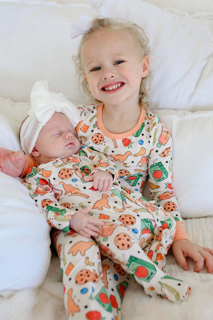 Smiling big sister holding a sleeping newborn wearing the Snack Attack Ruffle Bamboo Zipper Footie with a white bow headband, both children wearing coordinating Snack Attack print bamboo sleepwear