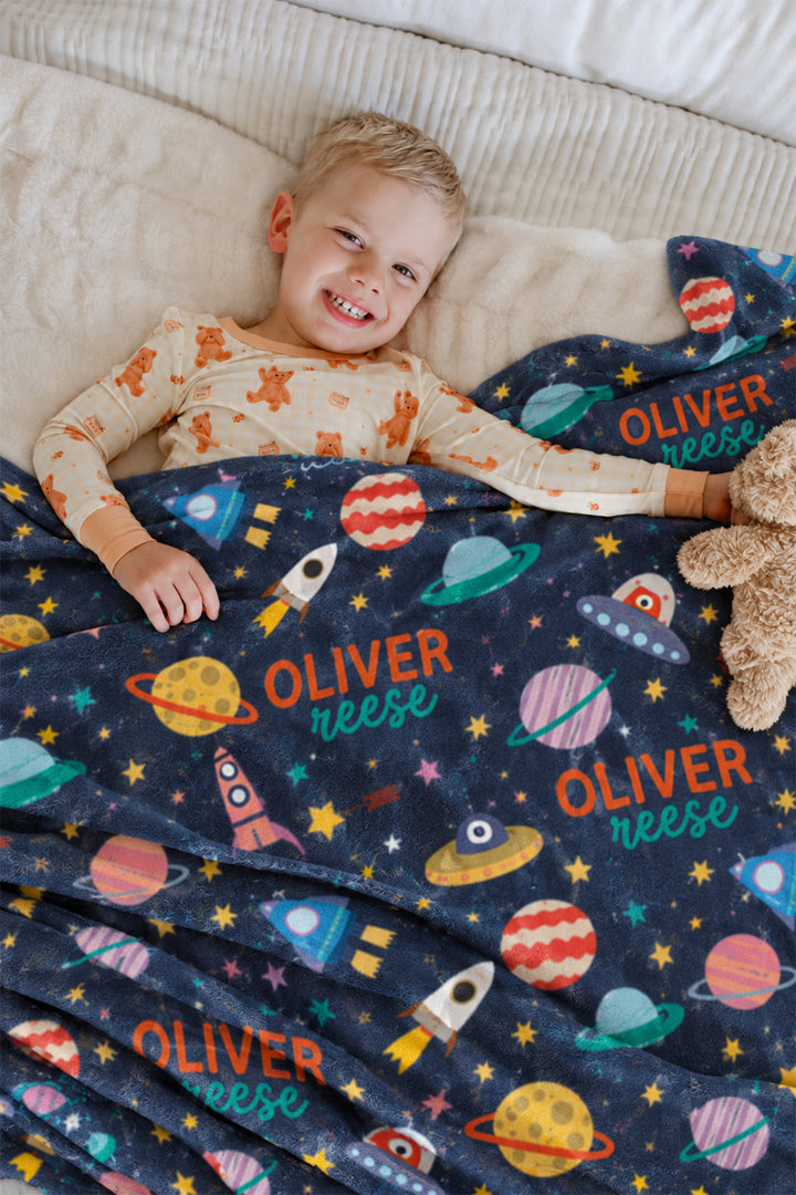 Smiling toddler lying in bed in bear-print pajamas, wrapped in a personalized Caden Lane Star Explorer blanket featuring planets, rockets, and a custom name