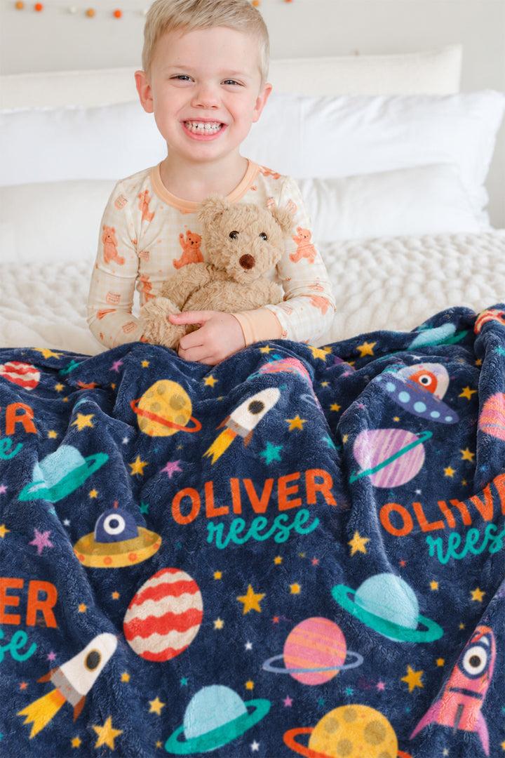 Toddler sitting on a bed hugging a teddy bear, behind a personalized Caden Lane Star Explorer blanket showing planets, UFOs, and custom name print