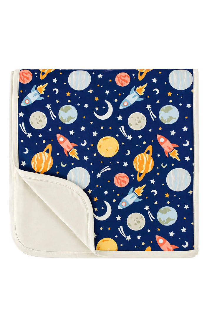Flat lay of a Caden Lane navy knit blanket in a Stellar Snuggles print featuring planets, rockets, and moons, with one corner folded to show the cream reverse.