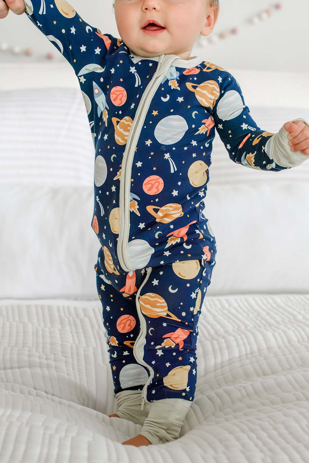 Baby standing on a bed with the two-way zipper open at one leg, wearing a Caden Lane navy bamboo romper in a Stellar Snuggles print featuring planets and rockets.