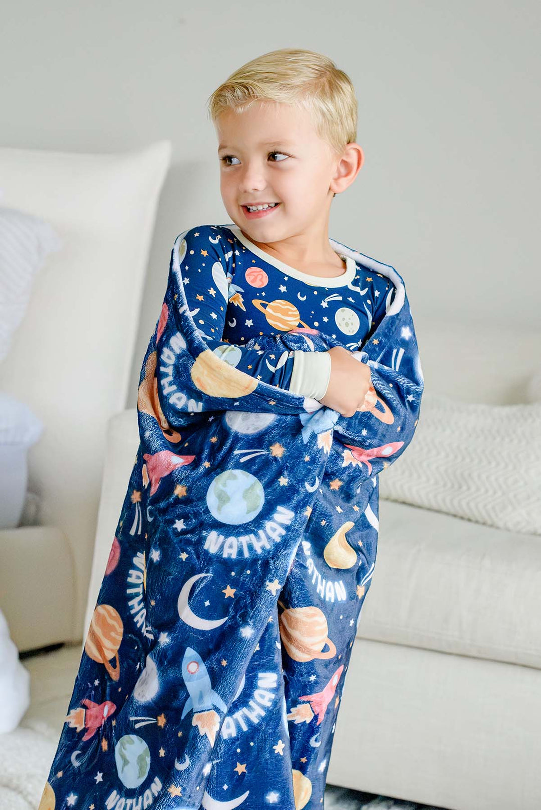 Smiling boy glancing to the side, wrapped in a Caden Lane navy plush blanket in a Stellar Snuggles print featuring planets and rockets, reading “Nathan,” paired with matching pajamas.