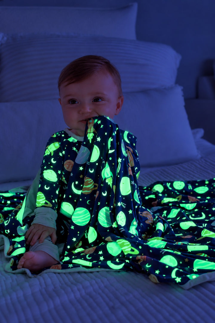 Baby sitting on a bed, chewing a glowing Caden Lane navy bamboo blanket in a Stellar Snuggles print featuring planets, moons, and rockets, paired with matching pajamas.