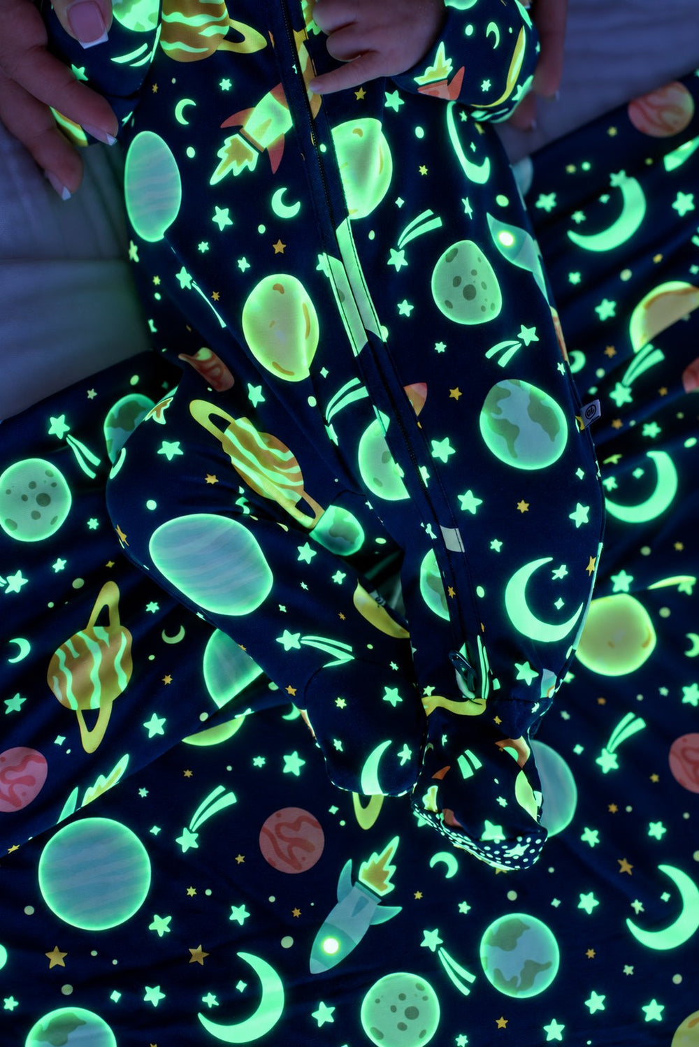 Close-up of a baby's legs and feet, wearing a glowing Caden Lane navy bamboo zipper footie in a Stellar Snuggles print featuring planets and moons, paired with a matching blanket.