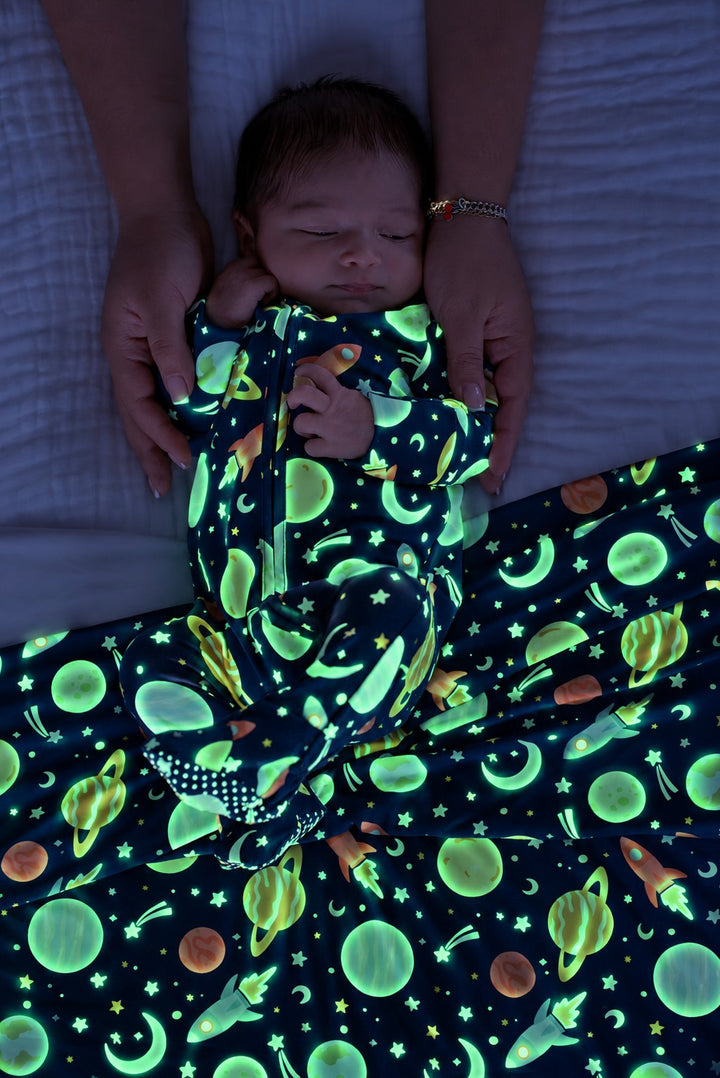 Sleeping newborn framed by adult hands, wearing a glowing Caden Lane navy bamboo zipper footie in a Stellar Snuggles print featuring planets and rockets, paired with a matching blanket.