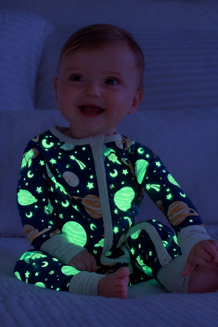 Laughing baby sitting against pillows on a bed, wearing a glowing Caden Lane navy bamboo zip romper in a Stellar Snuggles print featuring Saturn, crescent moons, and stars.