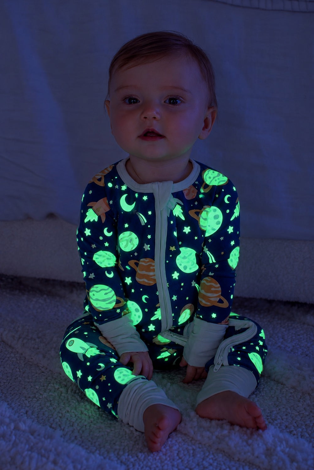 Wide-eyed baby sitting on a fluffy white rug, wearing a glowing Caden Lane navy bamboo zip romper in a Stellar Snuggles print featuring planets and shooting stars.