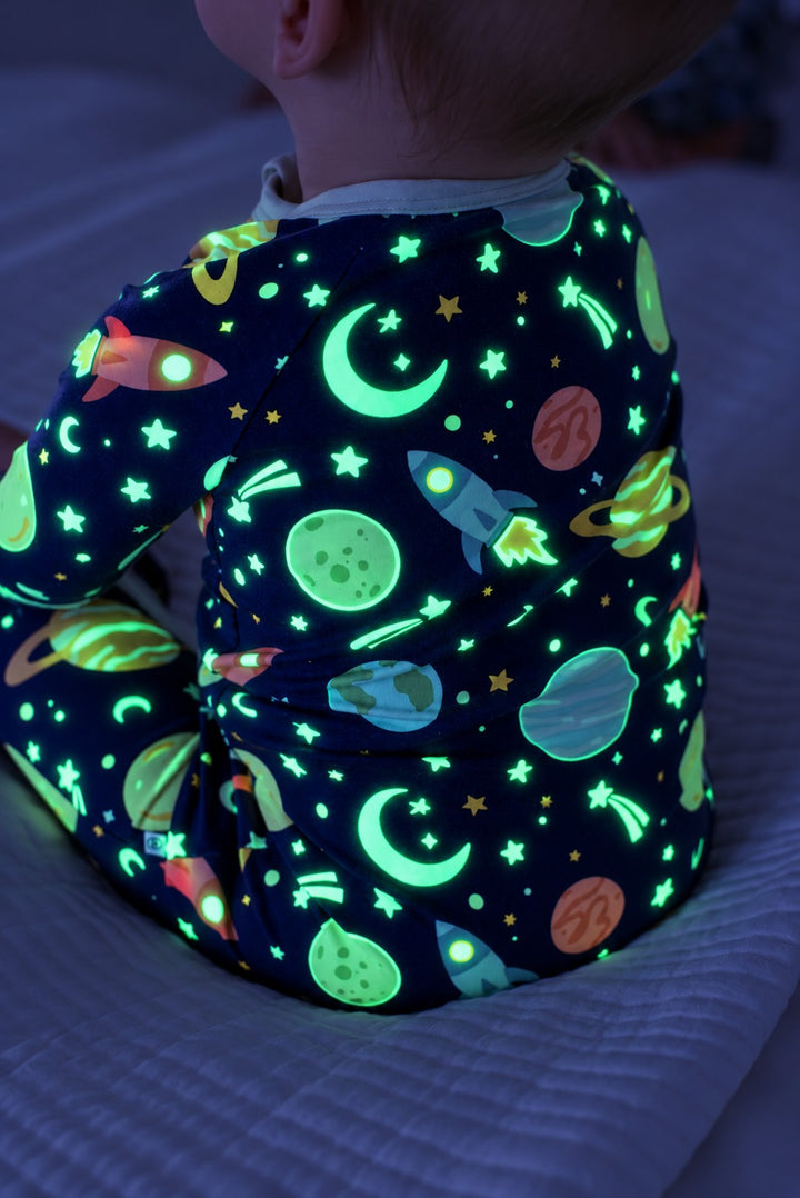 Back view of a baby sitting on a quilted bed, wearing a glowing Caden Lane navy bamboo zip romper in a Stellar Snuggles print featuring rockets, crescent moons, and planets.