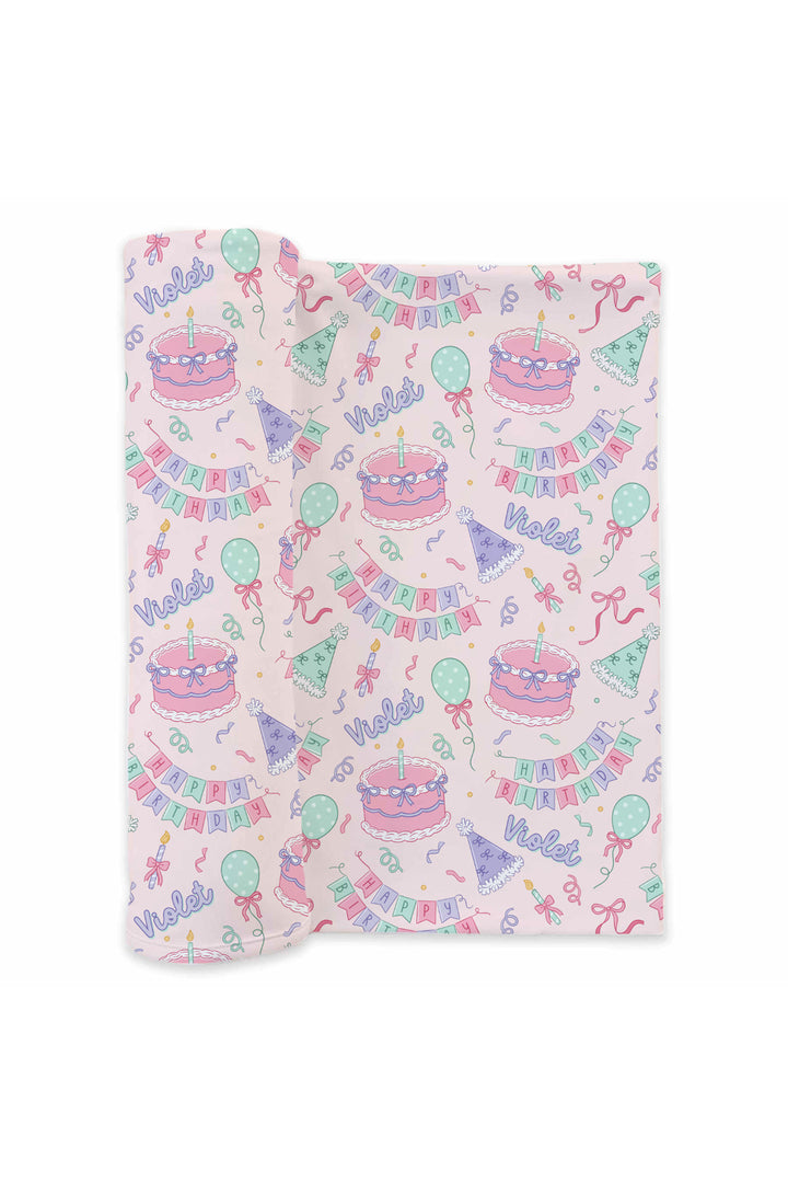 A Caden Lane folded baby swaddle with a light pink background and a colorful birthday print featuring cakes, balloons, banners, and happy birthday.