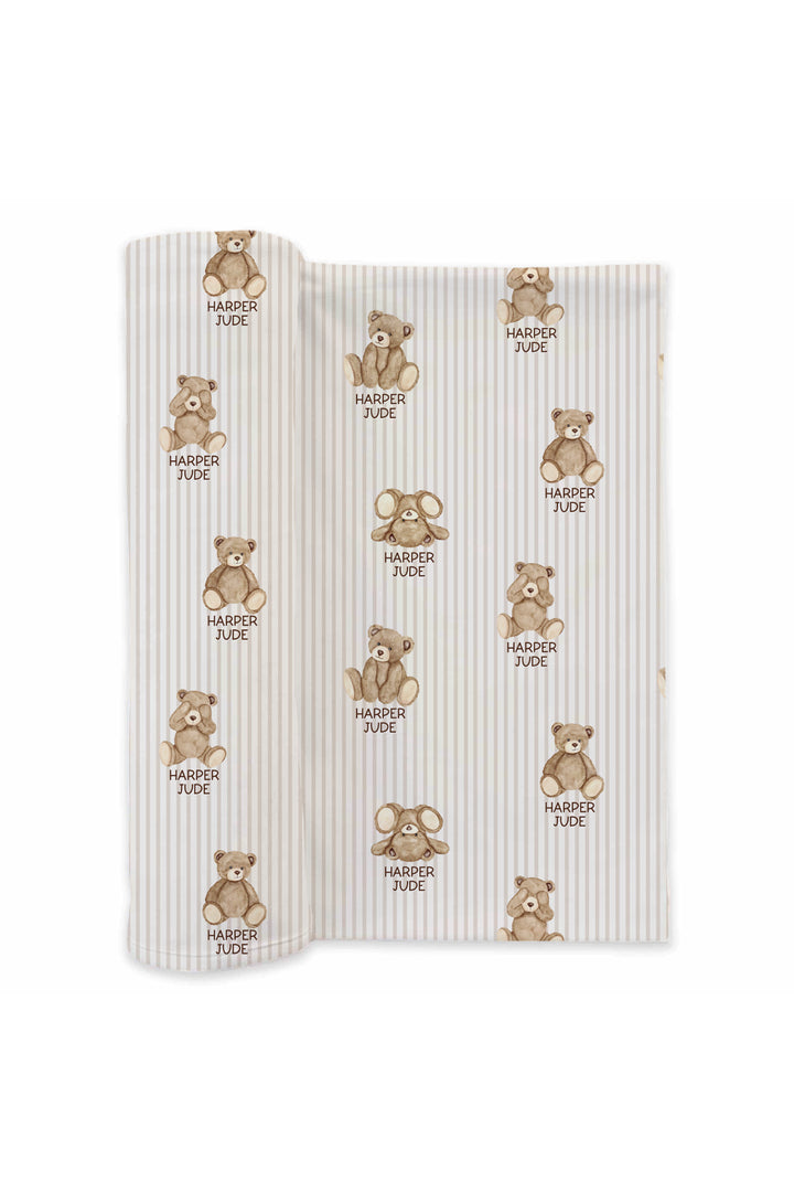 Flat lay of a partially unrolled Caden Lane cream knit swaddle in a Bear Stripe print featuring brown teddy bears and taupe pinstripes, reading "Harper Jude."