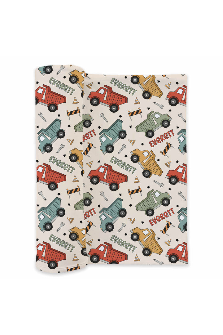 Flat lay of a partially rolled Caden Lane cream swaddle in a Busy Builder print featuring dump trucks, wrenches, and cones, reading "Everett" in red and sage.