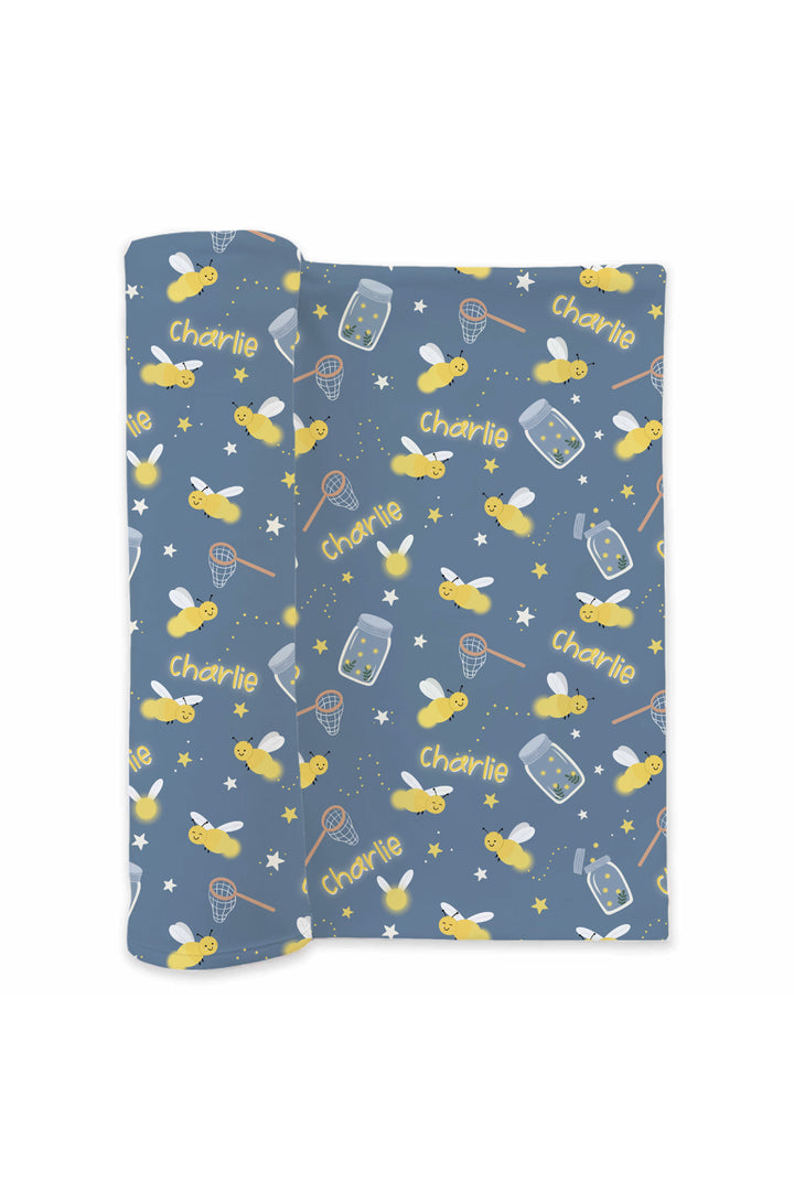 Product shot of a partially rolled Caden Lane blue personalized swaddle in a Chasing Fireflies print featuring fireflies and mason jars, repeating the name "Charlie."