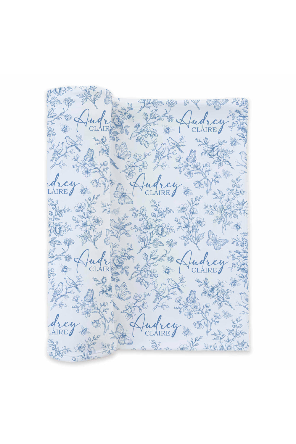 Flat lay of a partially unrolled Caden Lane blue and white knit swaddle in an Enchanted Meadow Toile print featuring florals, birds, and butterflies, reading "Audrey Claire."