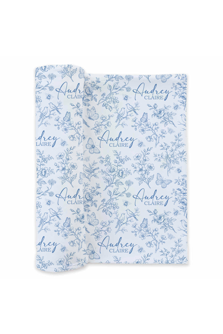 Flat lay of a partially unrolled Caden Lane blue and white knit swaddle in an Enchanted Meadow Toile print featuring florals, birds, and butterflies, reading "Audrey Claire."