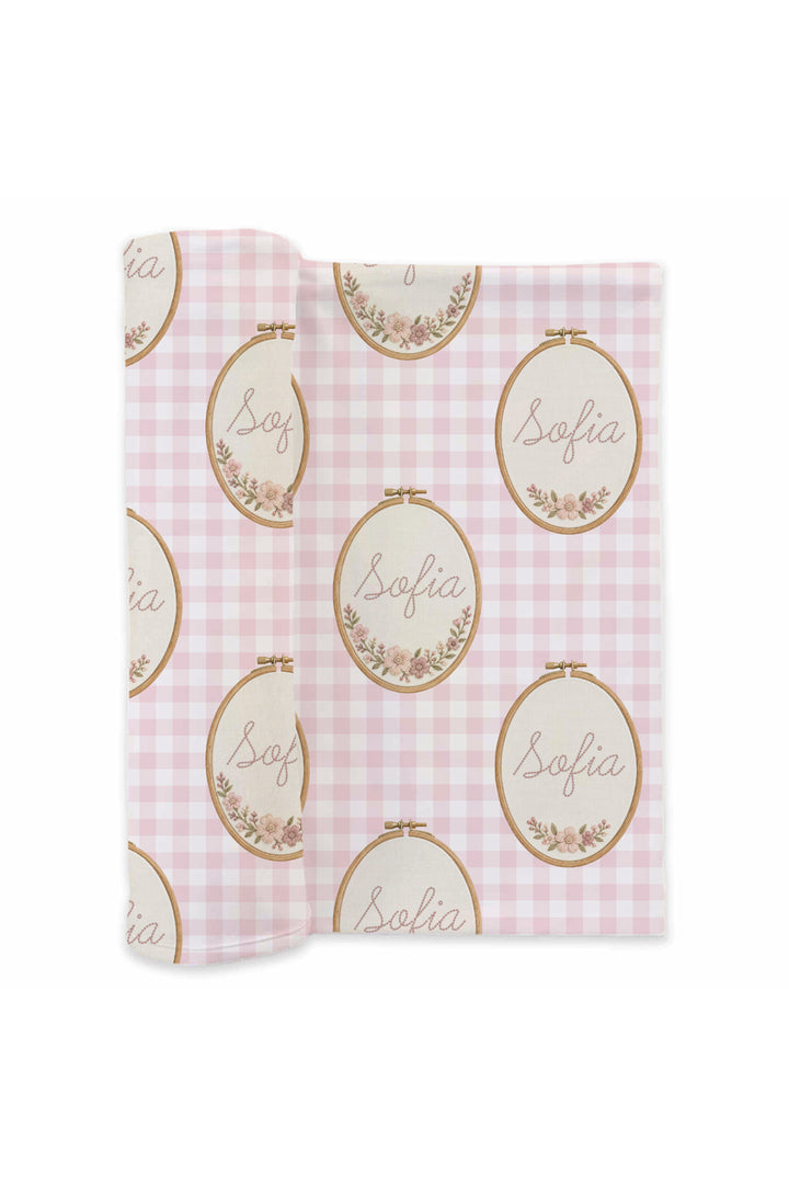 Flat lay of a partially unrolled Caden Lane pink knit swaddle in a Gingham Garden print featuring pink gingham and floral embroidery hoops, reading "Sofia."