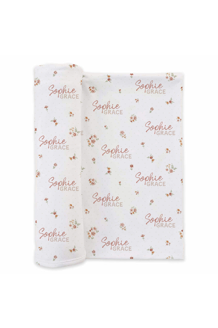 Flat lay of a partially unrolled Caden Lane white knit swaddle in an Heirloom Meadow print featuring tiny red rosebuds and leaves, reading "Sophie Grace."