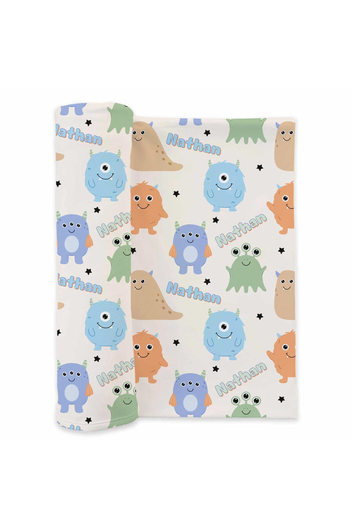 Product shot of Caden Lane personalized "Nathan" swaddle blanket rolled out, showing Little Monsters print with blue, purple, orange, tan, and green cartoon monsters and stars on a cream background