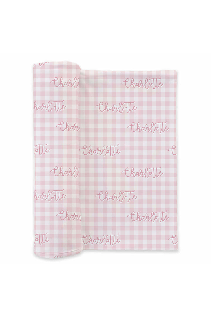 Flat lay of a partially unrolled Caden Lane pink knit swaddle in a Personalized Gingham print featuring pink and white checks, reading "Charlotte" in script.