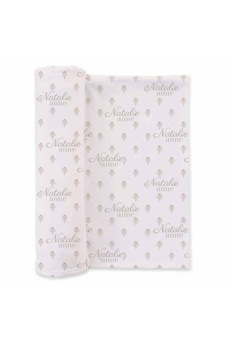 Flat lay of a partially unrolled Caden Lane white knit swaddle in a Vintage Classic print featuring small mauve floral sprigs, reading "Natalie Anne."