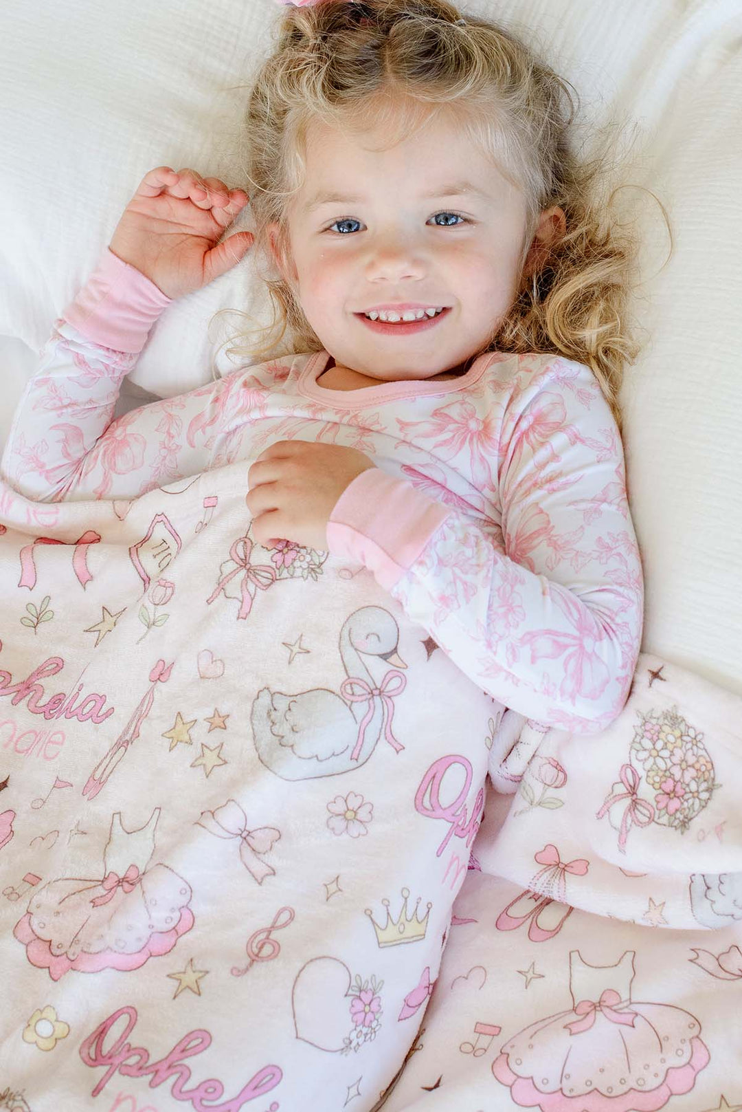 Smiling toddler lying on a bed wearing pink floral pajamas, snuggling with a personalized Caden Lane Swan Ballet blanket featuring swans, ballet slippers, and a custom name