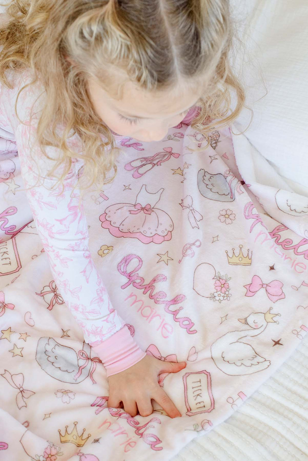 Close-up of a toddler leaning over a personalized Caden Lane Swan Ballet blanket, touching the fabric and showing the swan, ticket, and script name details