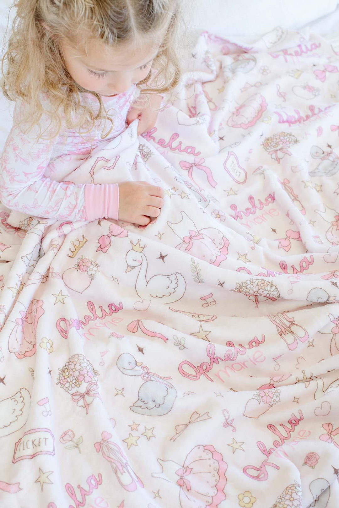 Toddler nestled in a rumpled personalized Caden Lane Swan Ballet blanket, showing swan, bow, and custom name print detail