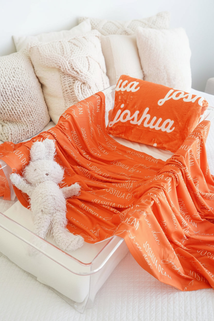 Orange blanket with 'Joshua' printed on it, accompanied by a white teddy bear and pillow.