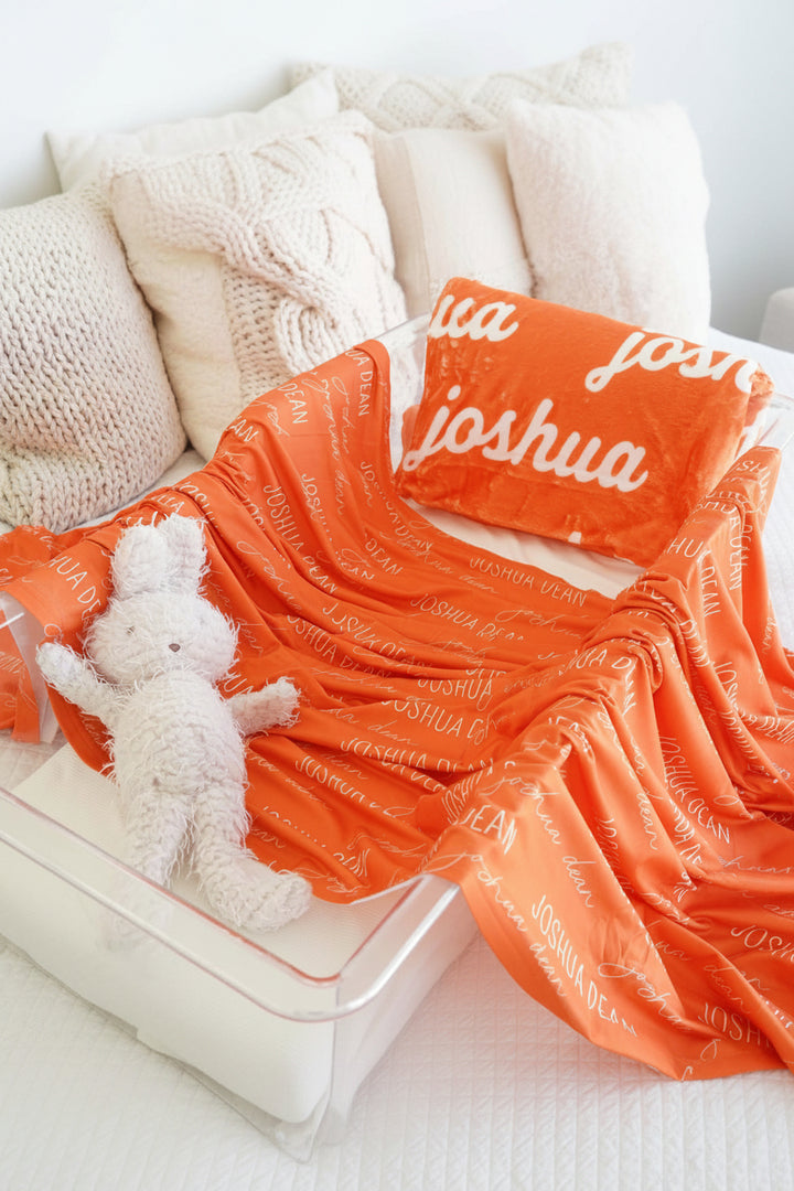 Orange blanket with 'Joshua' printed on it, accompanied by a white teddy bear and pillow.