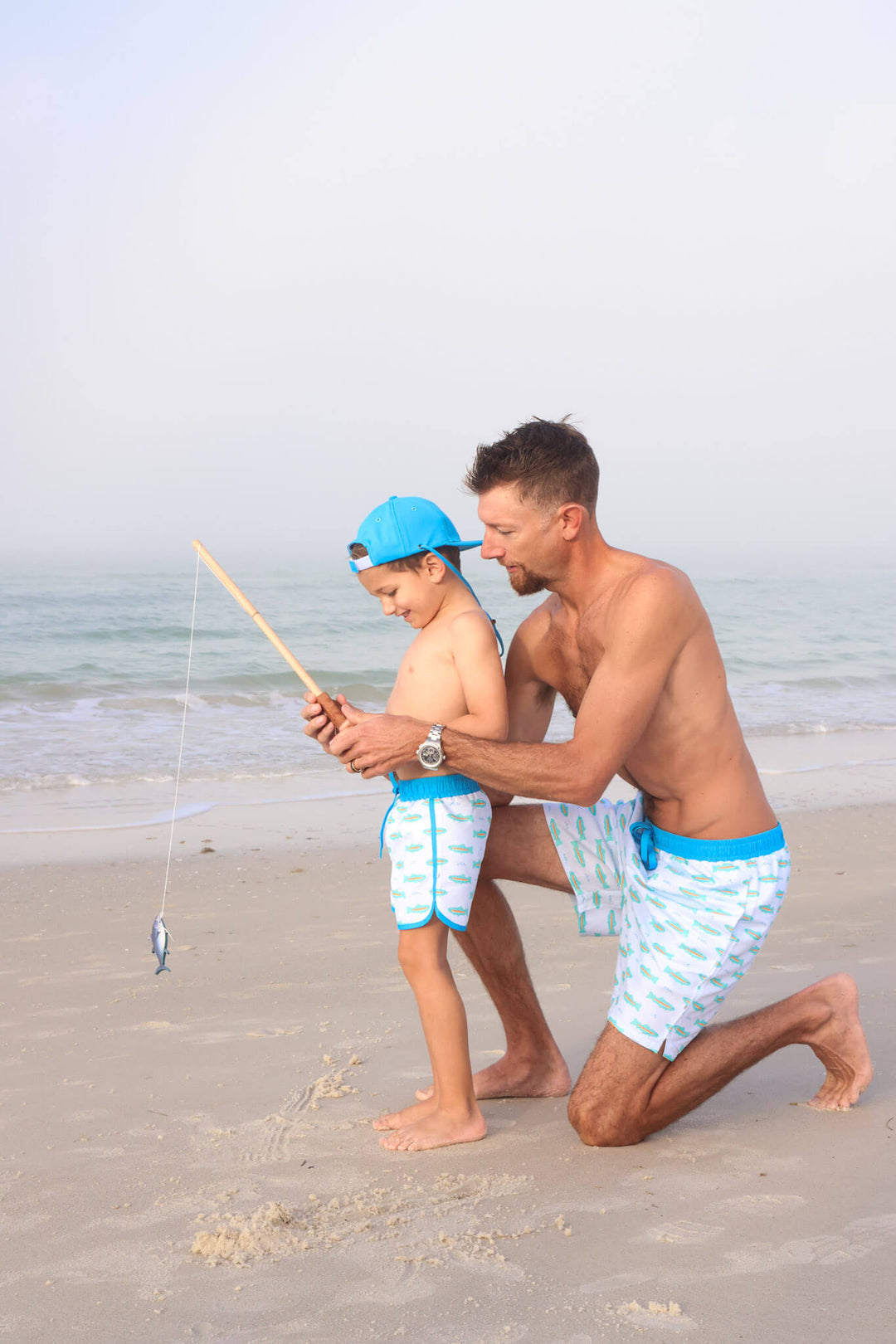 dad helping son fish on beach wearing matching trout swim trunks 
