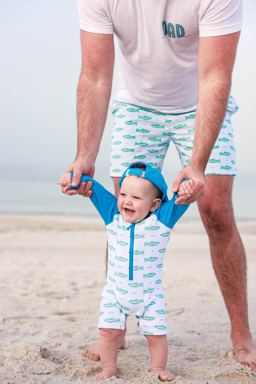 dad helping baby walk on beach in trout matching swim trunks and romper 