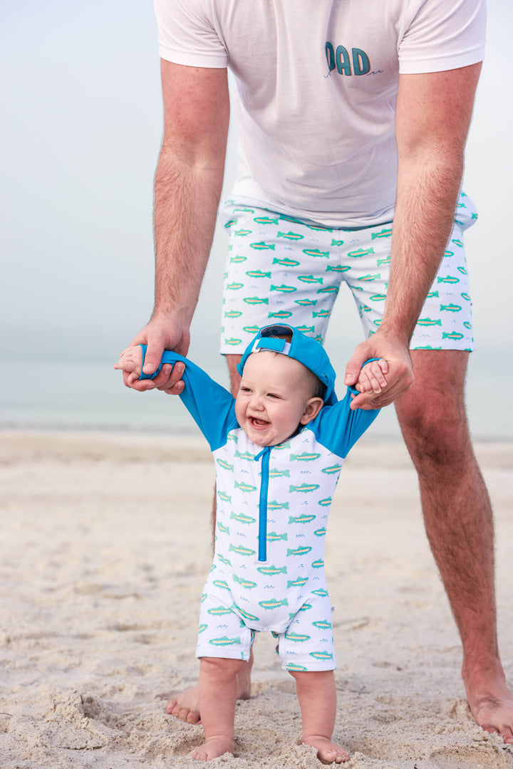 dad helping baby walk on beach wearing matching tiny trout swimsuits 