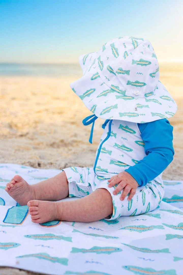 baby boy sitting on beach in tiny trout swim romper and hat 