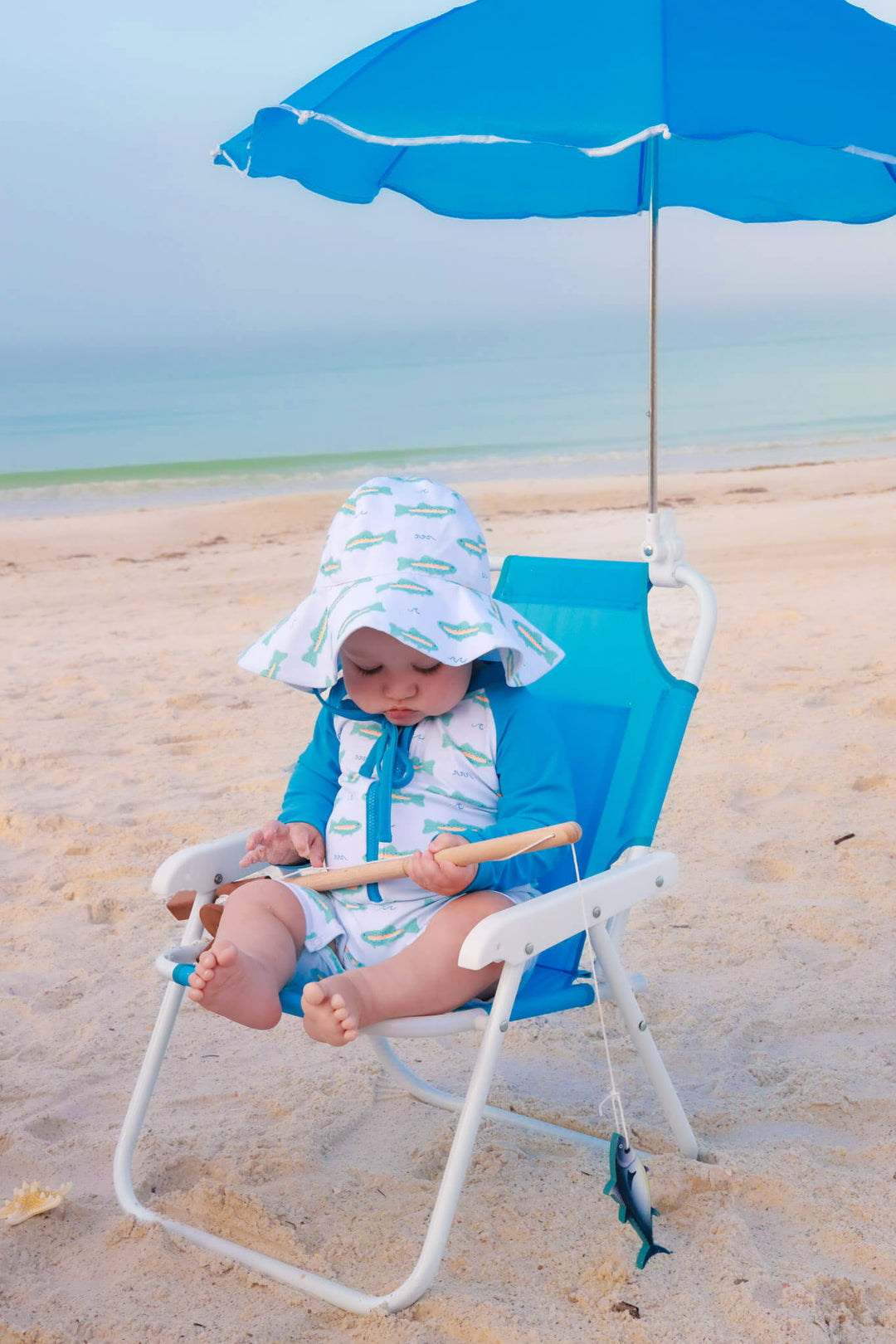 baby boy sitting in beach chair in tiny trout swim romper and matching hat 