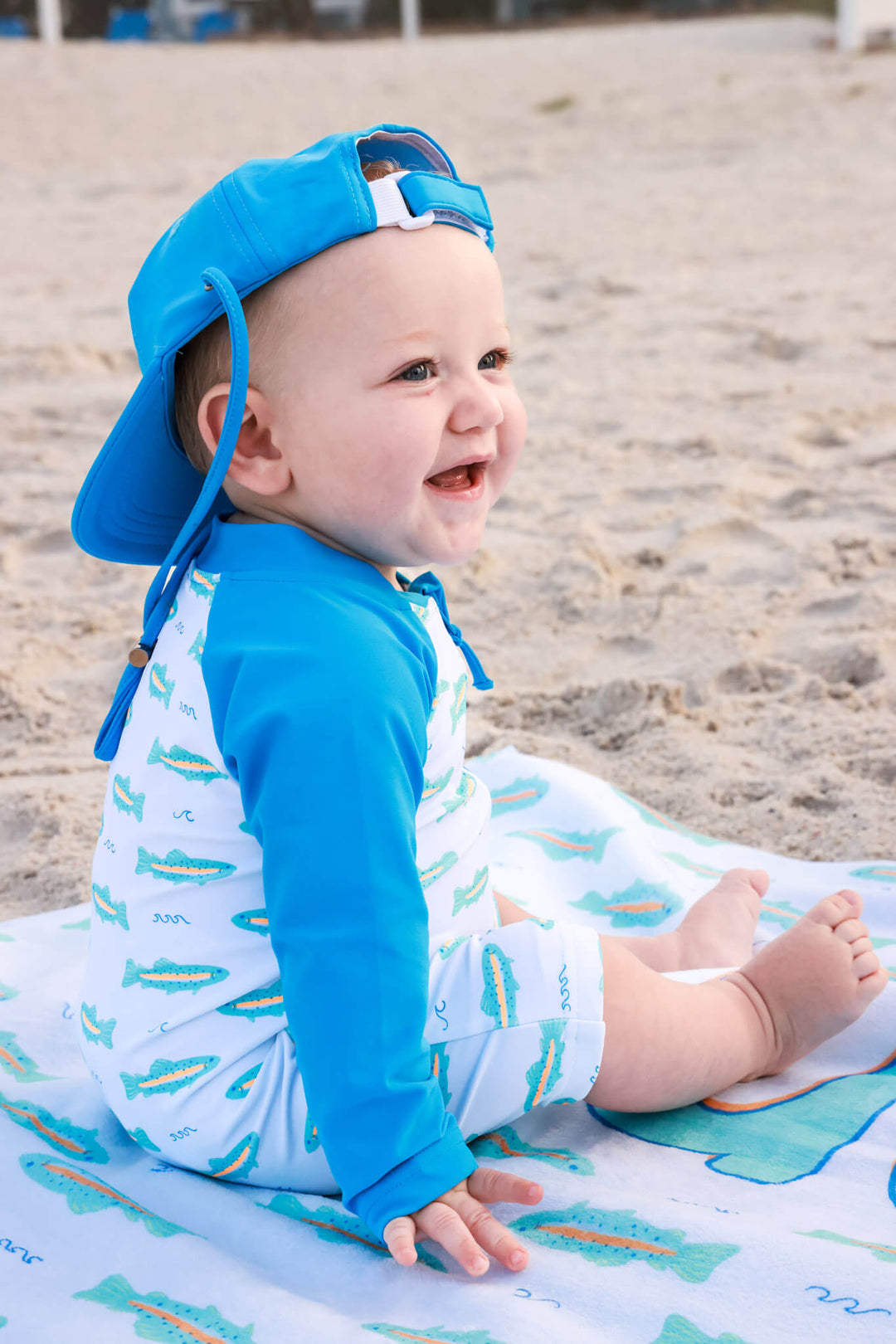 baby boy sitting on beach in tiny rout swim romper with bright blue surf hat 