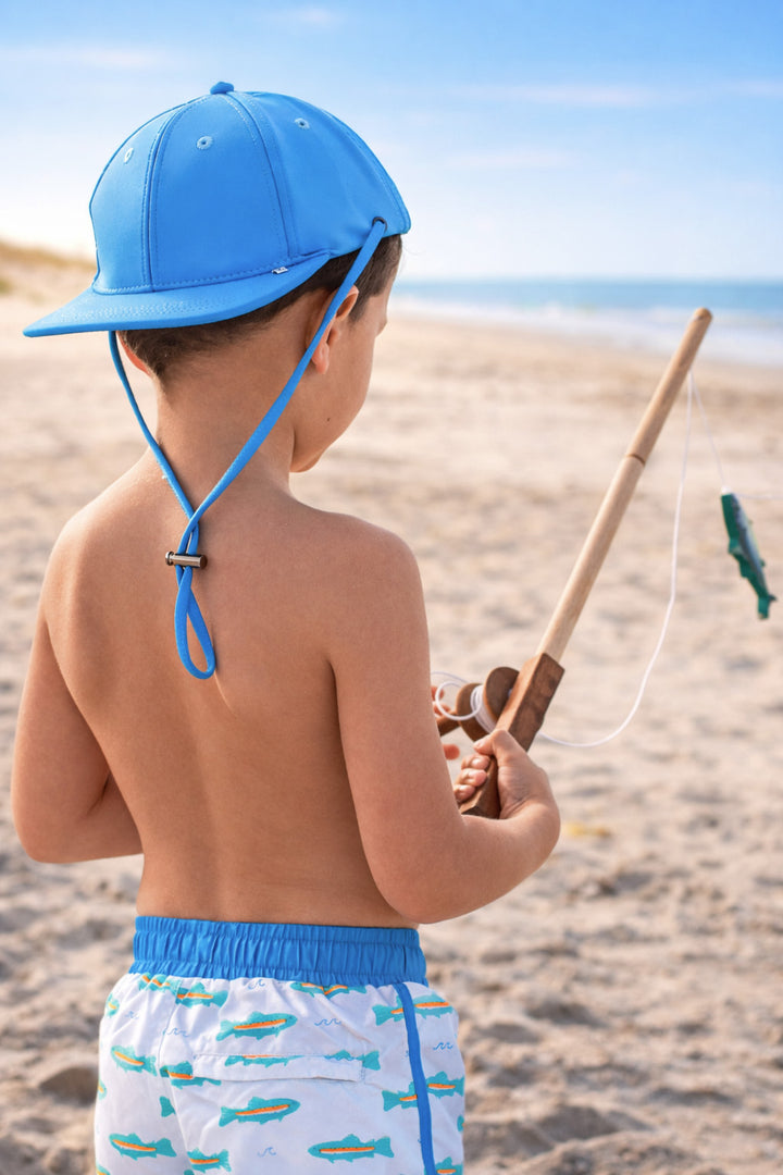 boy standing on beach with fishing pole in blue surf hat and tiny trout swim trunks 