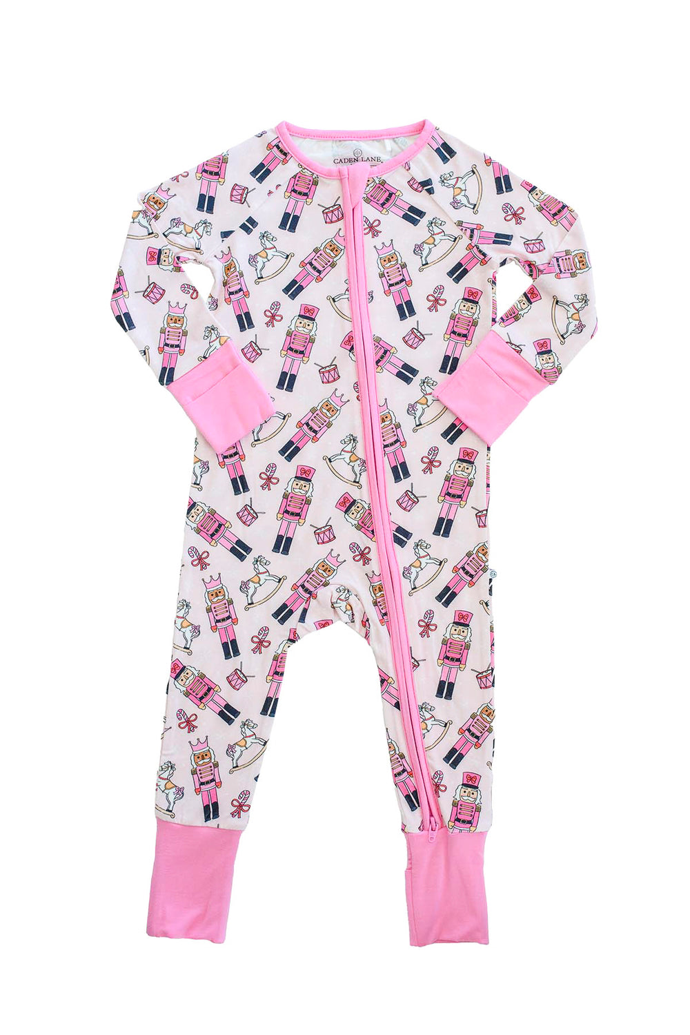 Flat lay of Caden Lane's pink toy soldier print zip-up pajamas, featuring nutcrackers and rocking horses, with pink cuffs and trim.