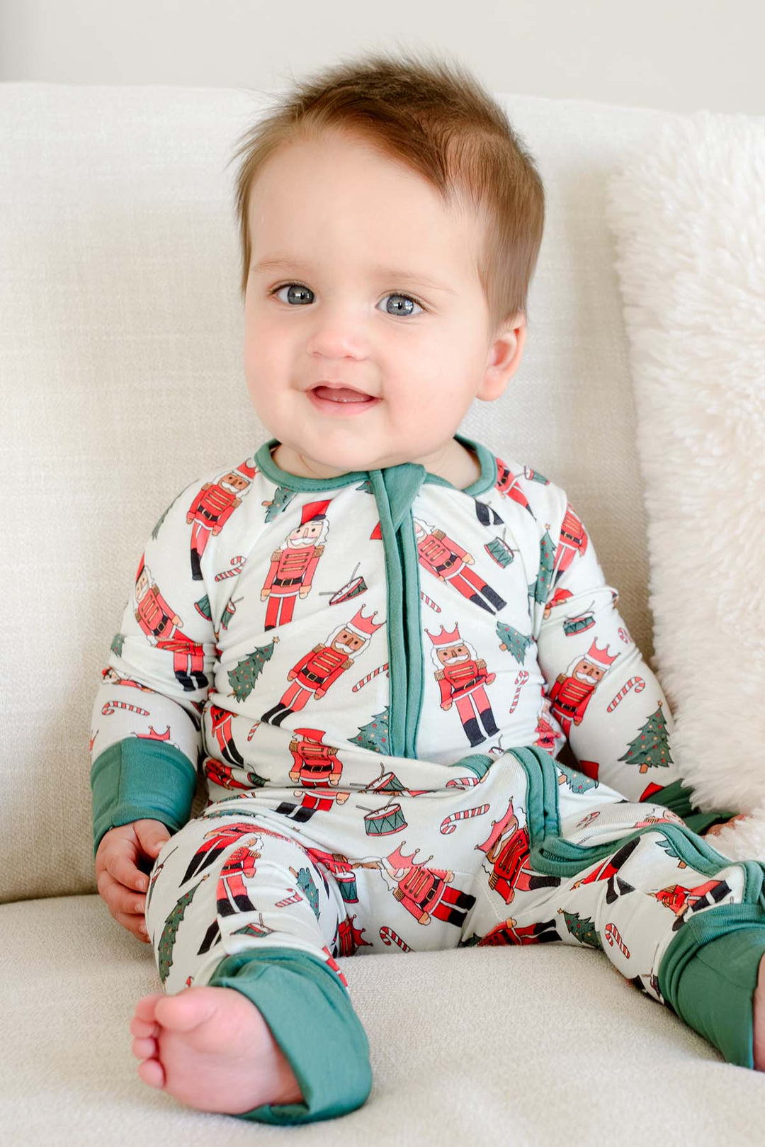 Baby boy smiling while wearing Caden Lane's white toy soldier print zip-up pajamas, featuring nutcrackers and Christmas trees, with green cuffs.