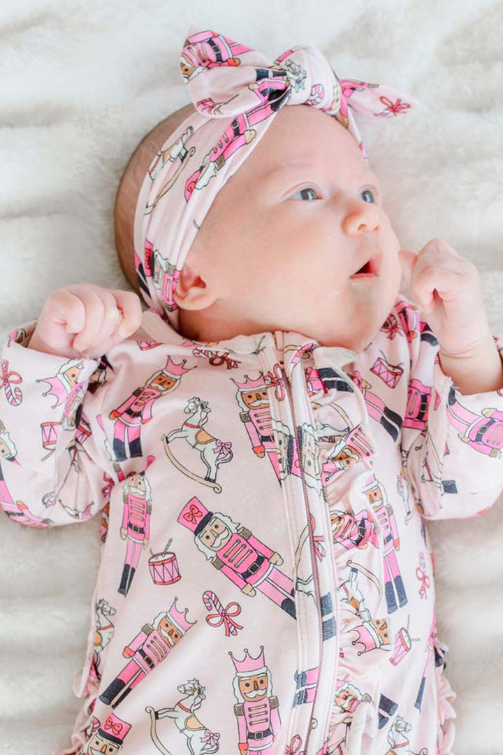 Newborn girl wearing Caden Lane's pink toy soldier print knot headband and matching ruffle zip-up pajamas, featuring nutcrackers and rocking horses.