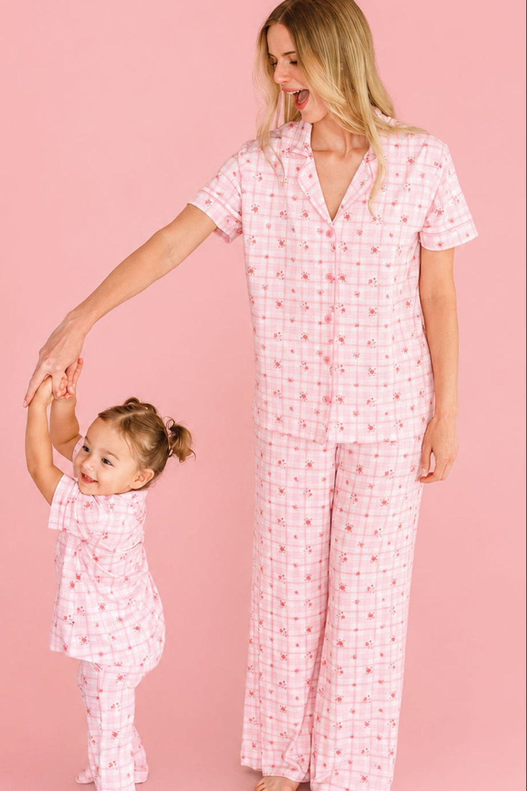 A woman and a young girl wearing Caden Lane matching pink patterned pajamas, with the woman holding the child's hand against a solid pink studio background.