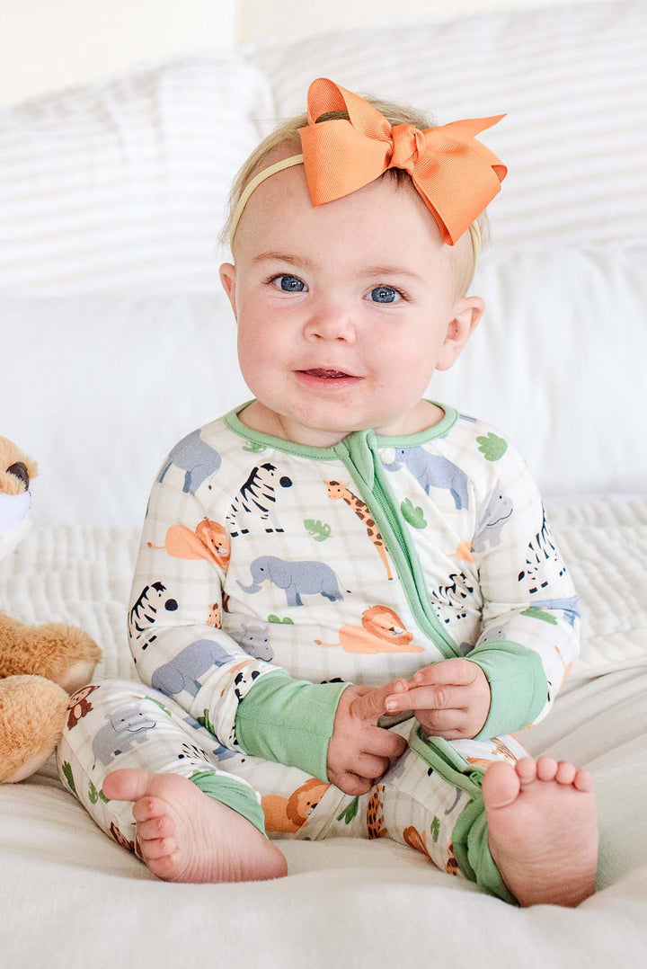 Smiling baby girl sitting on a bed in a Caden Lane convertible zip romper in a Wild Child print with sage green trim, paired with an orange bow headband and a lion plush.